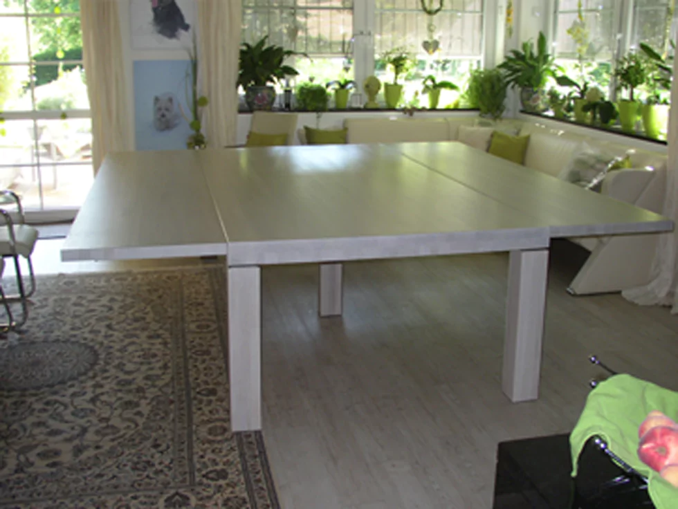 Ein weißer, ausklappbarer Tisch, der in einem Wohnzimmer steht. Im Hintergrund steht auch eine weiße Eckcouch und darüber stehen auf einem Vorsprung mehrere Pflanzen.