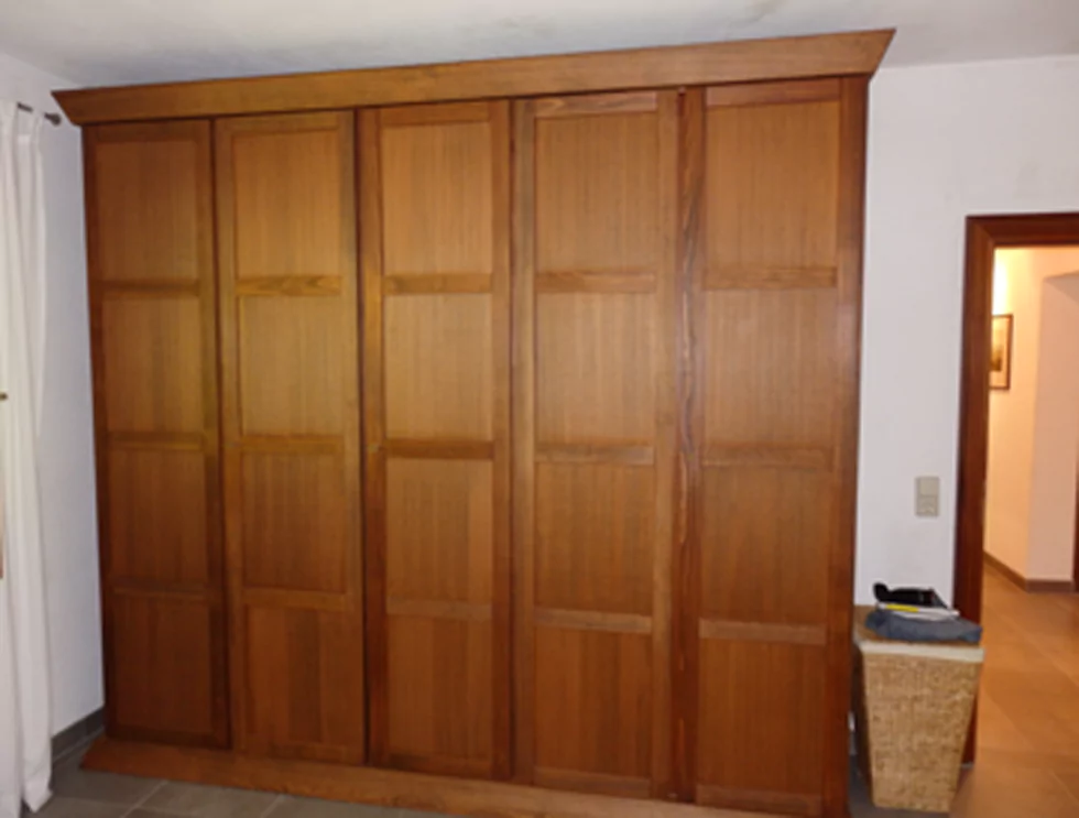 Ein brauner Schrank, der in einem Schlafzimmer steht. Rechts daneben steht ein brauner Flechtwäschekorb. Der Schrank reicht bis unter die Decke und hat 5 Türen zum Öffnen.
