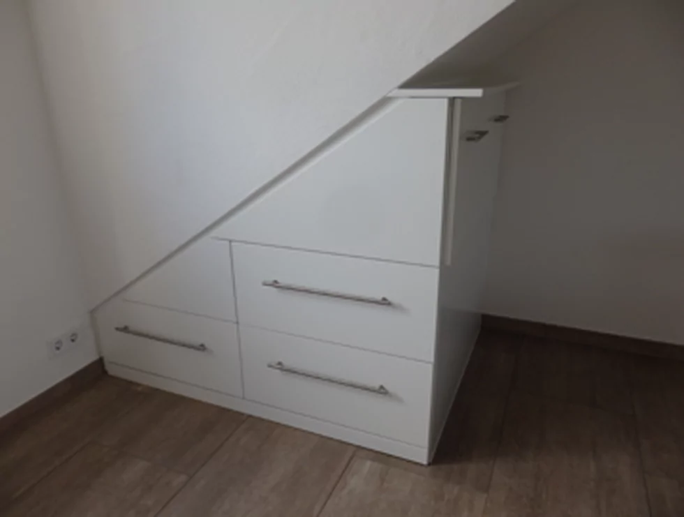 Ein weißer Schrank, der unter einem Dachvorsprung angebracht wurde. Dieser verläuft von links unten nach rechts oben. Der Schrank hat 4 Schubladen.