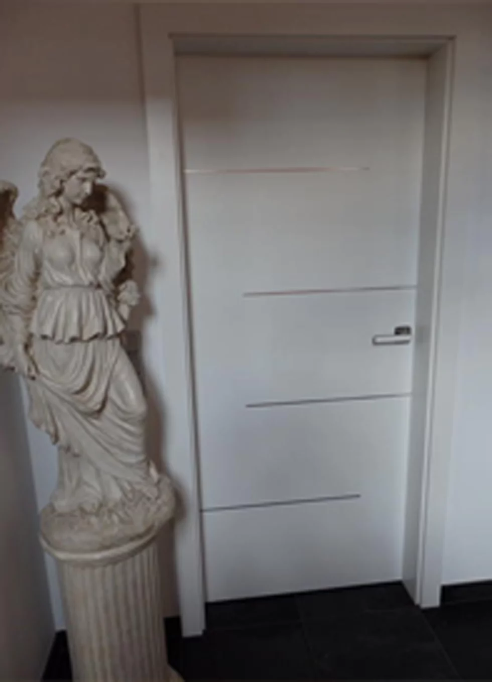 Eine weiße Tür, die mit vier silbernen Strichen waagerecht verteilt geschlossen in der Wand steht. Links daneben steht eine große weibliche Engelsstatue.
