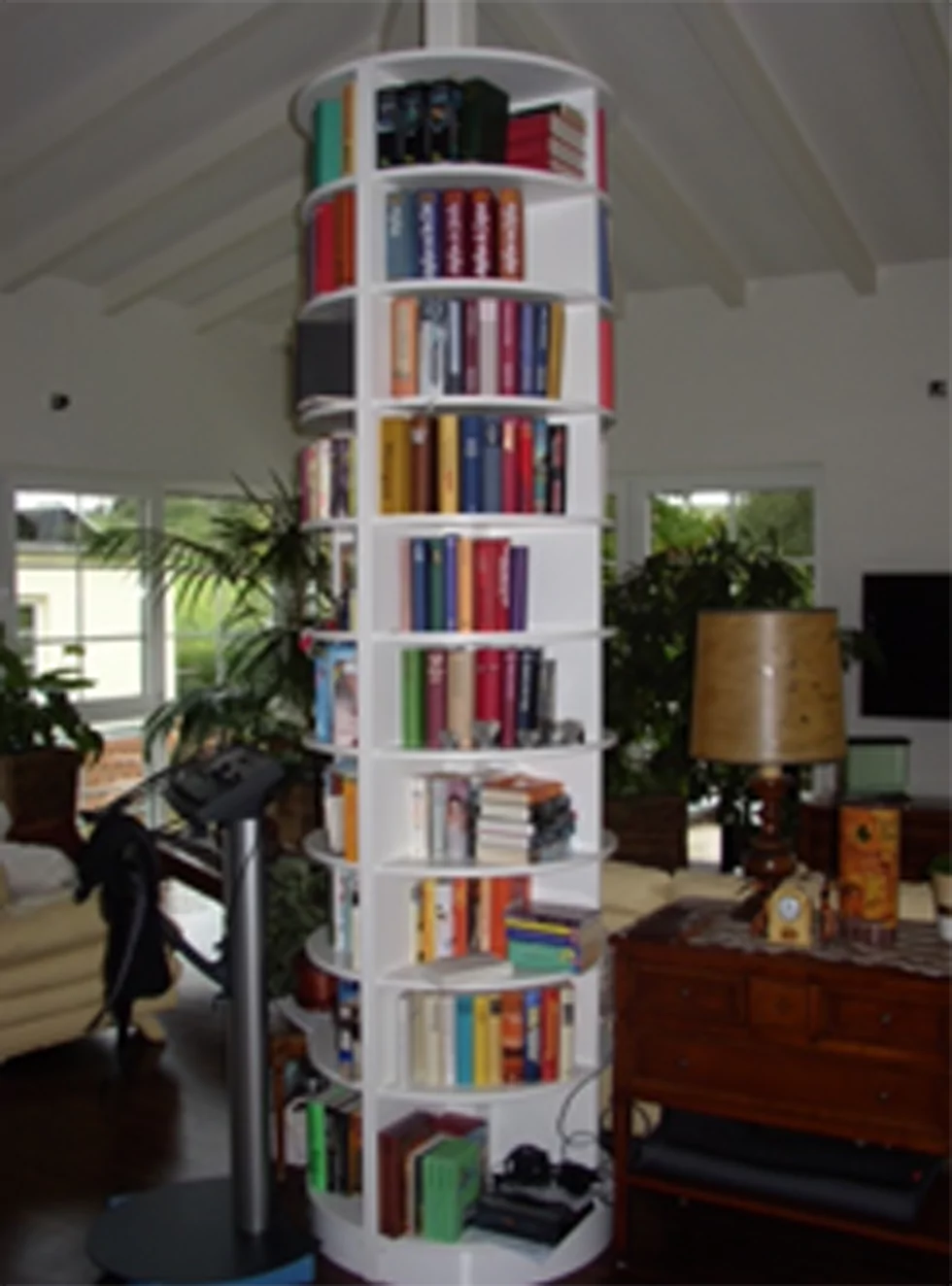 Ein weißes, rundes, drehbares Bücherregal steht in einem Wohnzimmer. Darin befinden sich zahlreiche Bücher. Im Hintergrund ist außerdem ein braunes Schränkchen mit einer gelben Lampe und Pflanzen zu sehen.