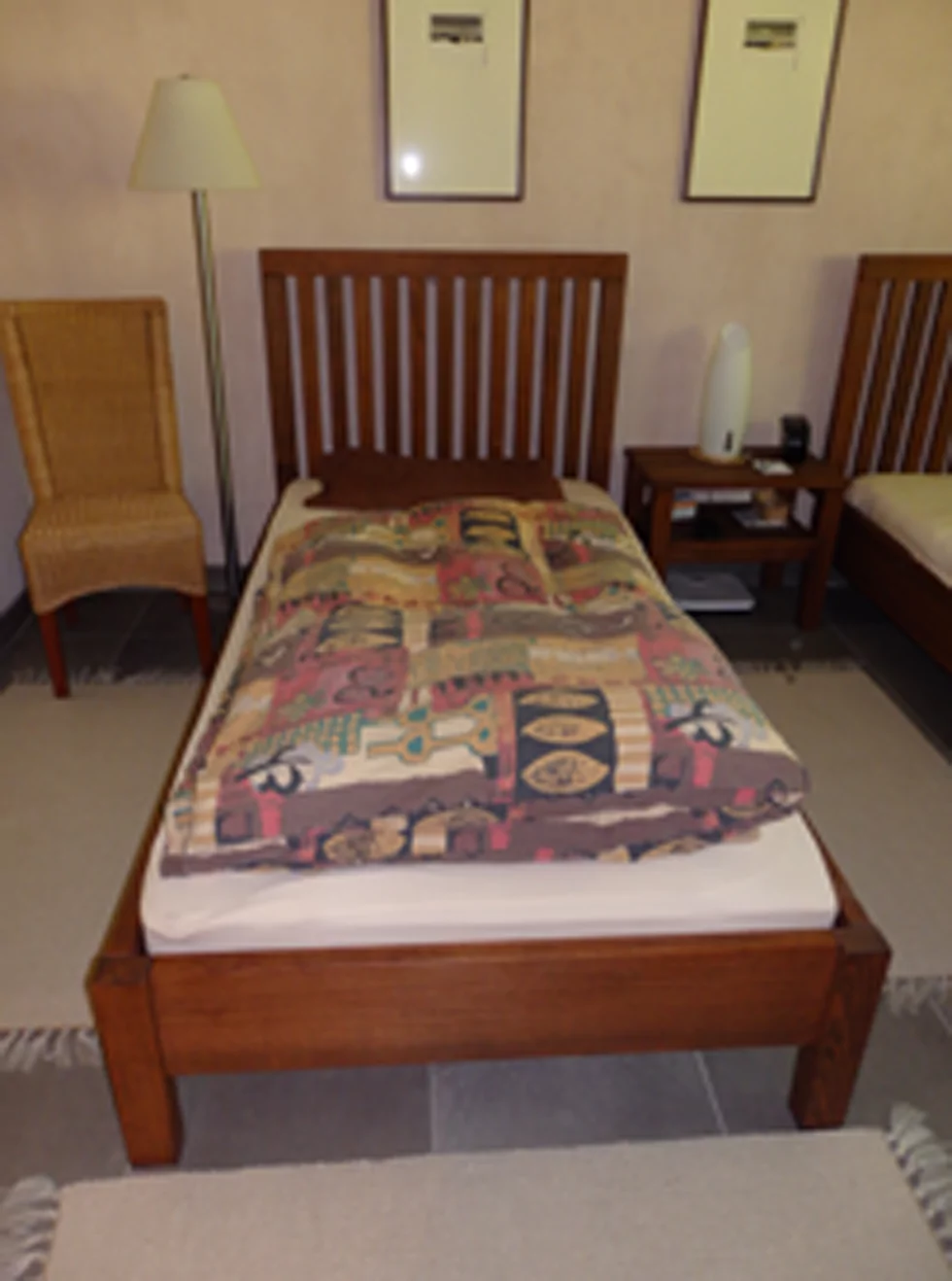 Ein braunes Bett mit weißem Bettlaken und bunter, mit Muster verzierter Bettdecke steht in einem Schlafzimmer. Links daneben steht ein brauner Stuhl und rechts daneben eine kleine braune Kommode.