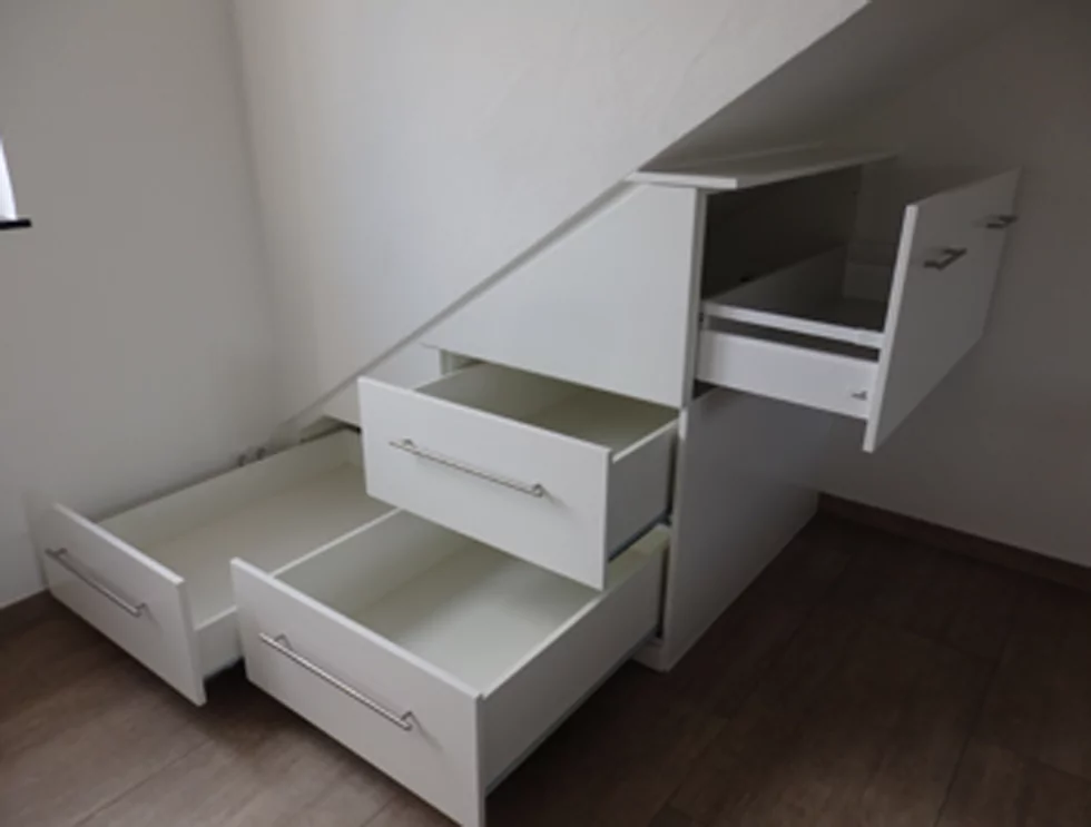 Ein weißer Schrank, der unter einem Dachvorsprung angebracht wurde. Dieser verläuft von links unten nach rechts oben. Der Schrank hat vier Schubladen. Es sind alle vier Schubladen geöffnet.